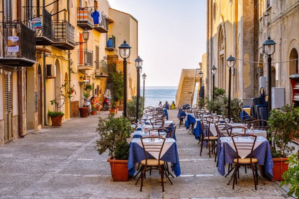 Tables set for dinner along a narrow Cefalù street in Sicily, opening onto the sea