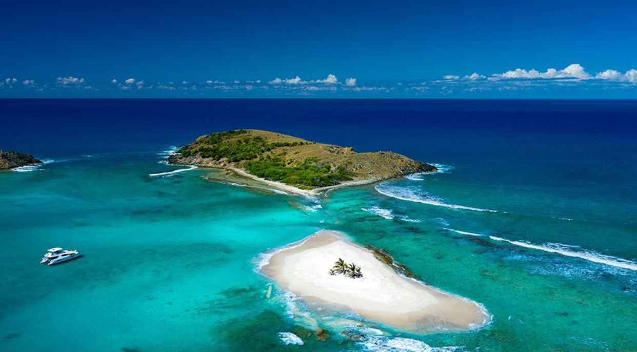Aerial view of a small Caribbean island ringed by turquoise shallows, a white sandbar and a single anchored catamaran