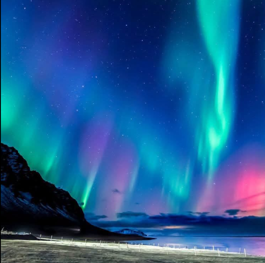 Green and violet aurora filling a star-lit sky above a dark coastal ridge and snow-covered shore